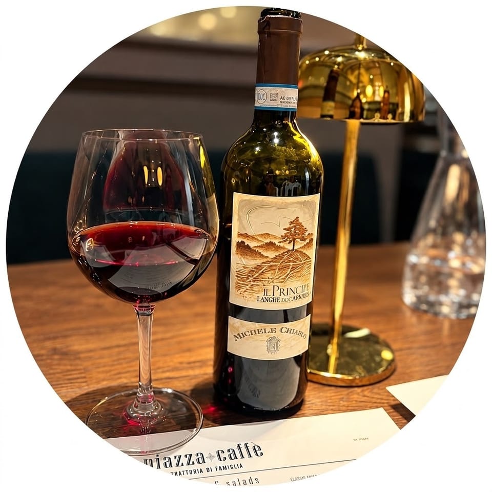 A glass of red wine and Michele Chiarlo bottle sit on a wooden table beside a lamp and Piazza Caffè menu.