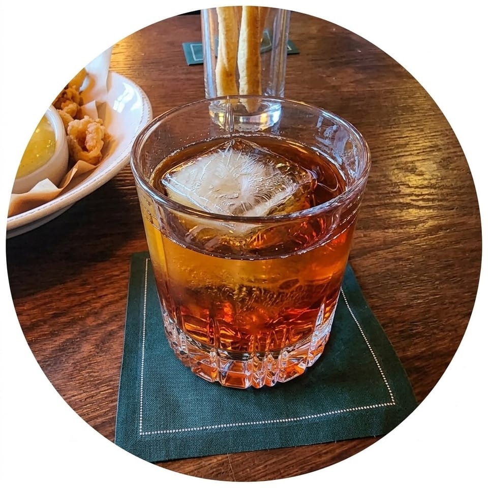Amber cocktail with a large ice cube on a green napkin, accompanied by fried shrimp and breadsticks on a wooden restaurant table.
