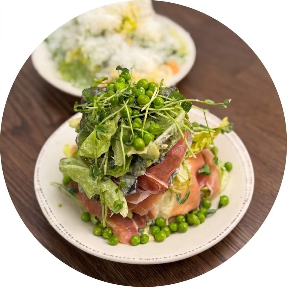 Stacked salad with smoked salmon, prosciutto, leafy greens, peas, and microgreens on a plate, with a blurred salad bowl behind.
